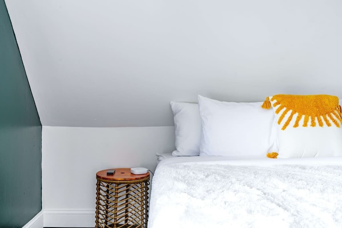 Een netjes opgemaakt bed met witte beddengoed en een geel dekentje, een klein rond rieten bijzettafeltje met een wit object erop, en een effen witte muur.