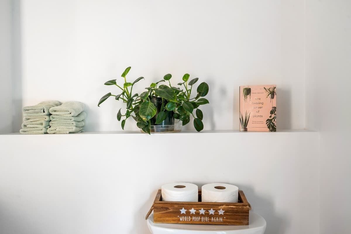 A bathroom shelf with a stack of folded towels, a potted plant, a book titled 'The Healing Power of Wildflowers', and a wooden box with toilet paper rolls.