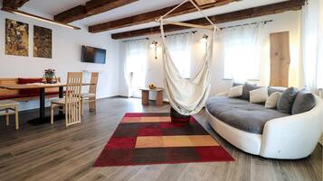 A modern living room with a sofa, a dining table with chairs, a hanging chair, and a TV mounted on the wall.