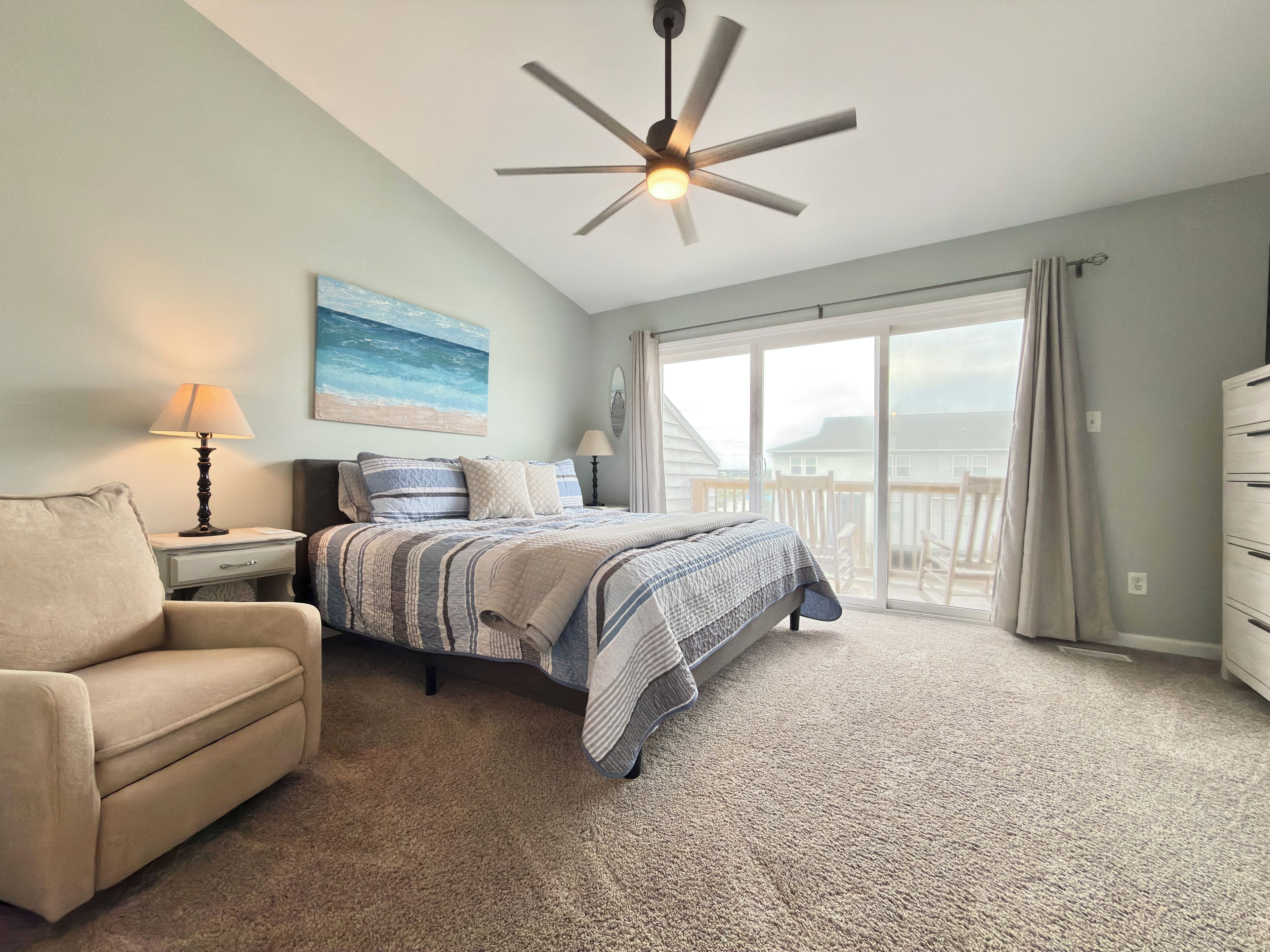 A bedroom with a bed, a chair, a lamp, a ceiling fan, a painting, and a view through a large window.