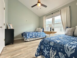 Une chambre avec deux lits, un ventilateur de plafond, une fenêtre avec des rideaux et une table de chevet avec une lampe.
