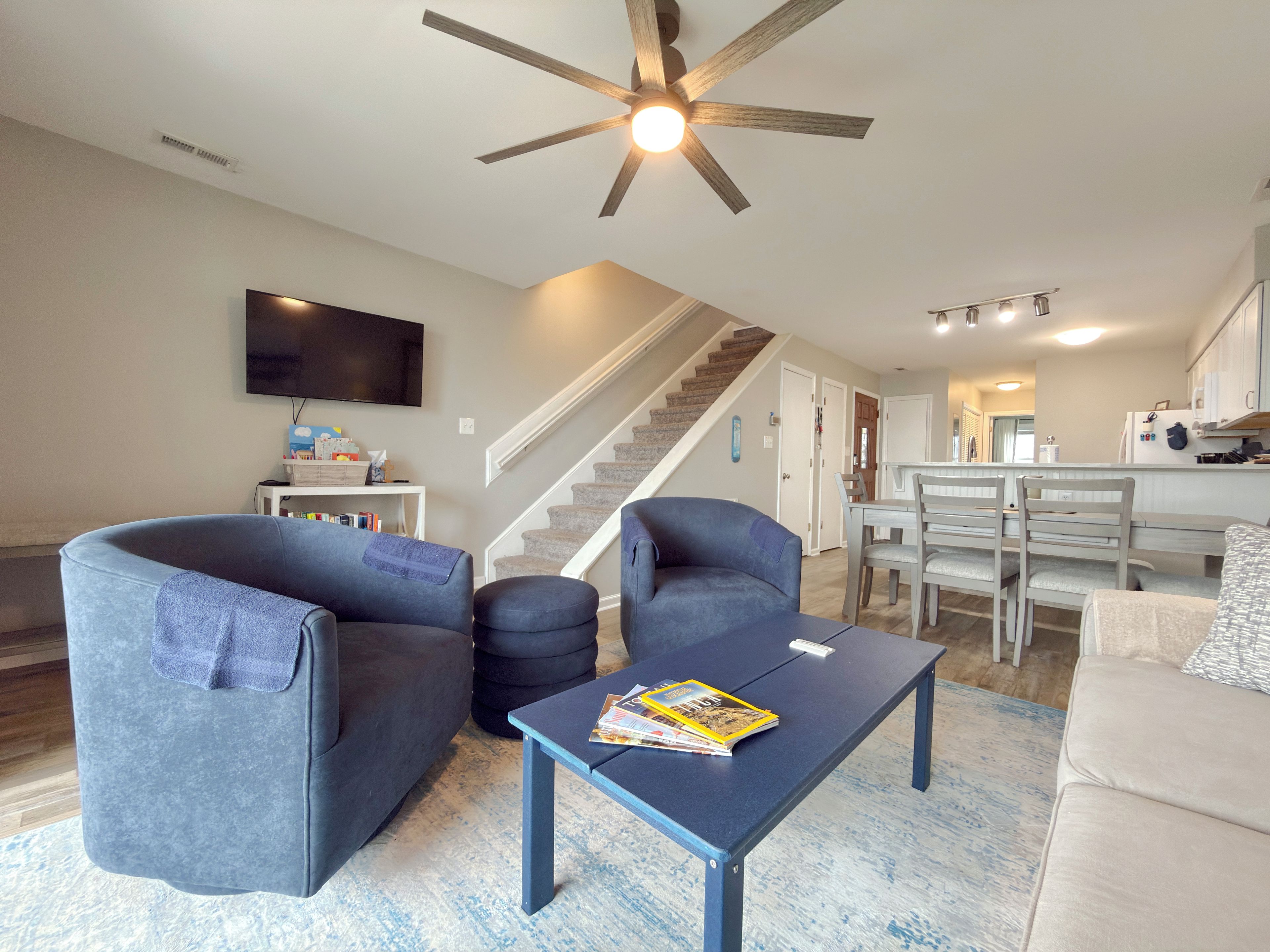 A modern living room with a flat-screen TV, a ceiling fan, and a staircase leading to the kitchen.