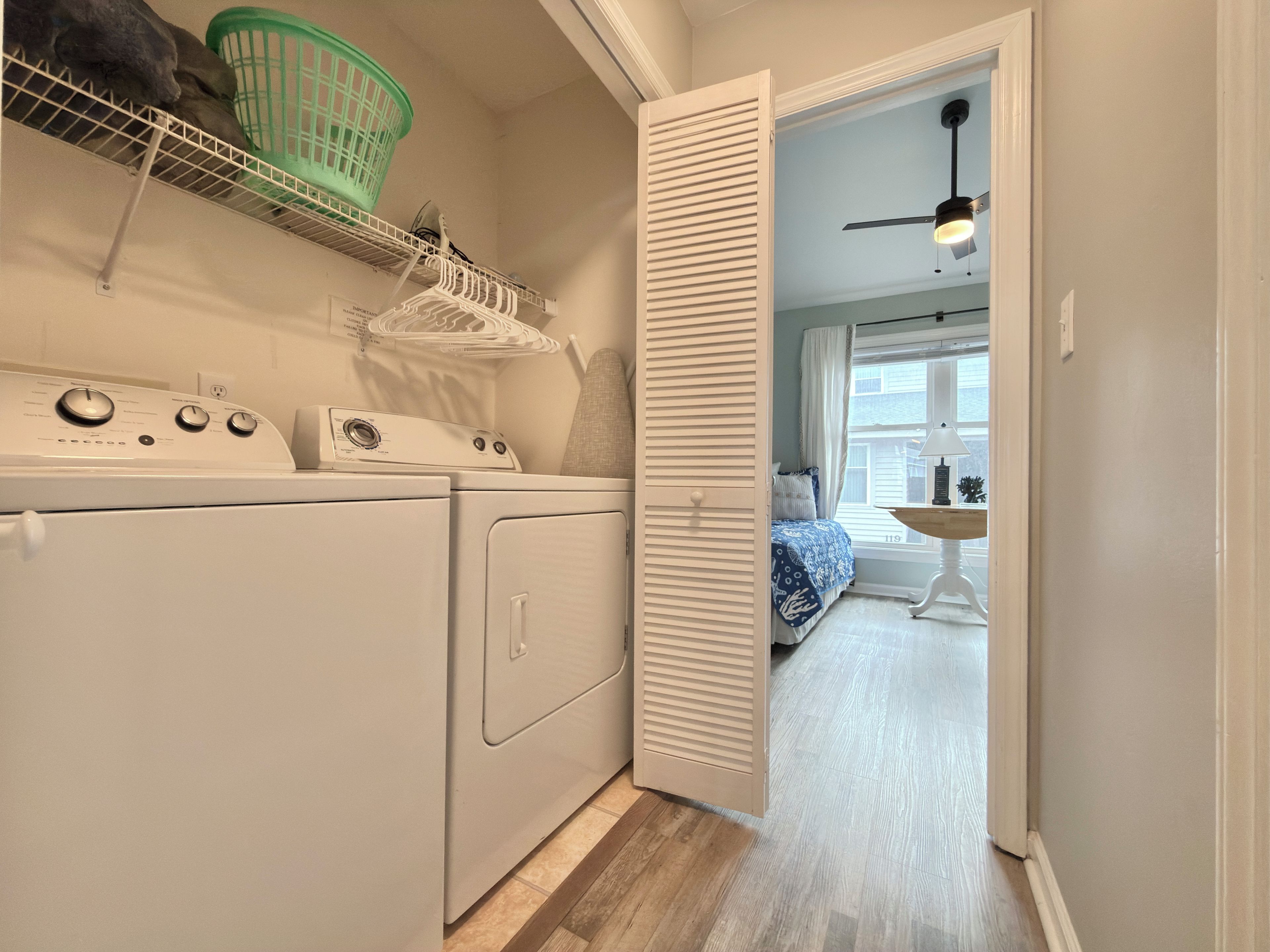 A laundry room with stacked washer and dryer units, a green laundry basket on a shelf, and a door leading to a bedroom with a bed and a nightstand.