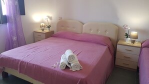A neatly made bed with pink bedding and rolled towels, two bedside tables with lamps and small plants, and a window with purple curtains.