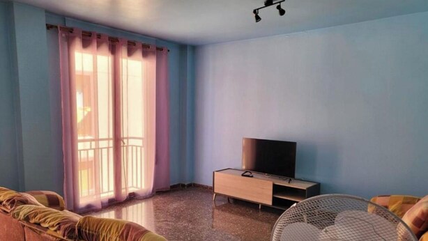 Een woonkamer met een bank, een televisie, een ventilator en een raam met gordijnen.