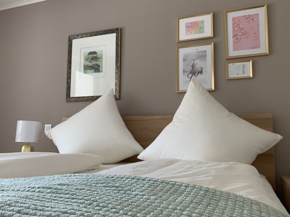 A neatly made bed with white pillows and a green blanket, a bedside lamp, and framed artwork on the wall.