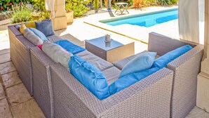 A wicker sofa with blue cushions and a small table with a candle, set on a patio with a pool in the background.