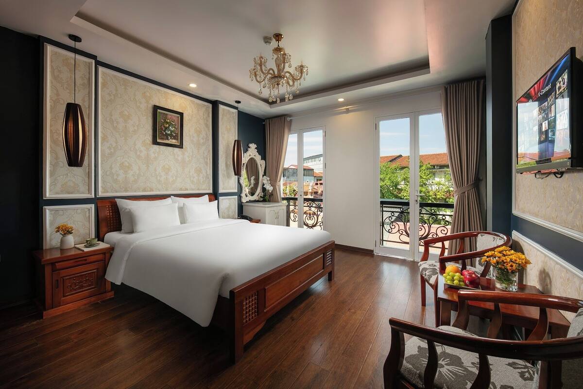 A hotel room with a large bed, wooden floor, a chandelier, a dining area with a table and chairs, and a balcony with a view of the city.