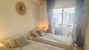 A bedroom with two beds, a small bedside table with a lamp, a window with curtains, and a view of the outside balcony.