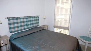 Una cama individual con cabecera de cuadros, una mesita de noche con lámpara y una ventana con vistas a edificios.