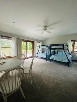Una habitación con literas, ventilador de techo, una mesa con sillas y ventanas grandes.
