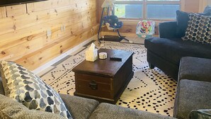 A cozy cabin interior with a wooden interior, a large TV mounted on the wall, a sofa, a coffee table, and a window with a view of trees.