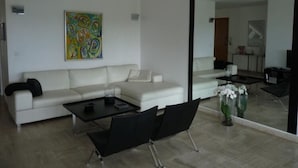 Un salon moderne avec un canapé blanc, une table basse noire et un fauteuil noir. Un tableau abstrait aux couleurs vives orne le mur, et un grand miroir reflète l’ensemble de la pièce.