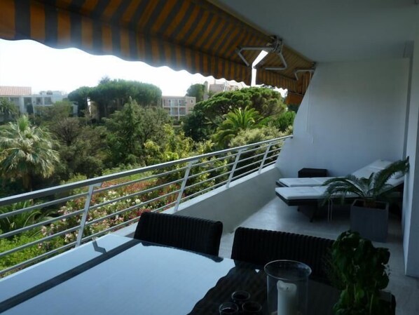 A balcony with a table and chairs, a railing, and a view of a lush garden and buildings.