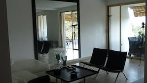 Un salon moderne avec un canapé blanc, une table basse noire et un grand miroir. Il y a un balcon avec une chaise et un trépied.