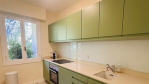 A kitchen with green cabinets, a white countertop, a window, a sink, and a trash can.