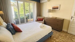 A hotel room with a large bed, a desk, a chair, a dresser, and a window with a view of greenery.