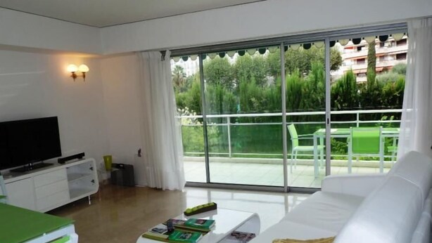 A modern living room with a white sofa, a flat-screen TV, and a coffee table with magazines. A balcony with a glass door is visible through the window. - Three bedroom, two bathroom apartment in Cannes with large terrace - 880 (Cannes)