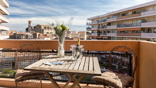 A balcony with a wooden table, two chairs, a vase with flowers, and a bottle of champagne. - Modern 3BR/3BA cannes apt walk to Palais - 2000 (Cannes)