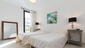 A bedroom with two beds, a bedside table with a lamp, a painting of flamingos, and a window with curtains. - Modern 3BR/3BA cannes apt walk to Palais - 2000 (Cannes)
