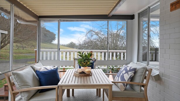 Eine überdachte Terrasse mit Holztisch und -stühlen, einem Blumentopf und Blick auf einen weißen Zaun und Bäume.