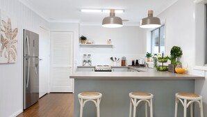 Eine moderne Küche mit weißen Schränken, einem Edelstahlkühlschrank, einem Gasherd und einer großen Kochinsel mit Barhocker. Über der Kochinsel befinden sich Pendelleuchten und ein Fenster sorgt für natürliches Licht.
