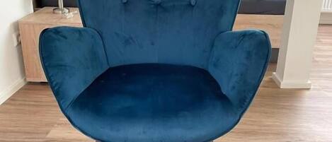 A dark blue upholstered armchair with tufted backrest and wooden legs, positioned in a room with wooden flooring and a small side table with a lamp.