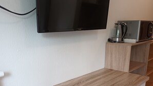 Een moderne kamer met een houten bureau, een grijze stoel, een flatscreen televisie aan de muur en een koffiezetapparaat op een plank.