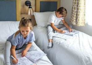 Deux enfants sont en train de dessiner sur des draps dans une chambre qui possède une tête de lit en bois, une lampe fixée au mur et un rideau à motifs de girafes.