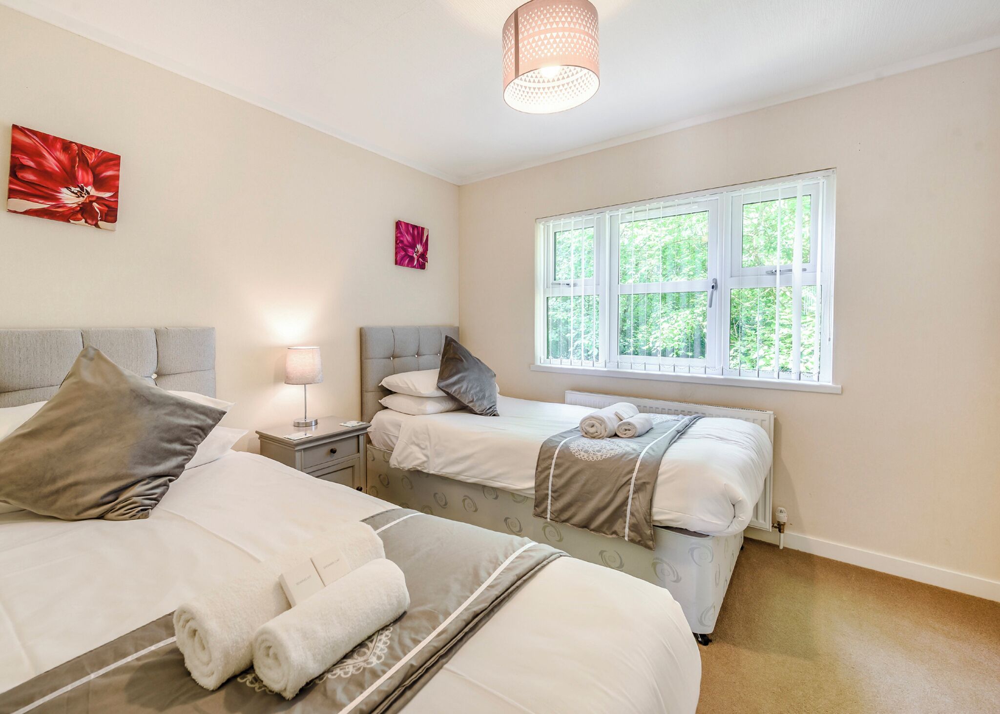 A bedroom with two beds, a bedside table with a lamp, and a window with a view of greenery.