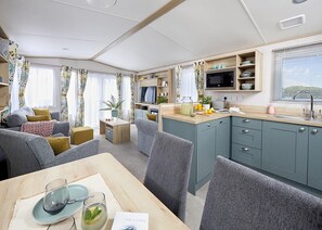 A compact living space with a dining area, kitchen, and seating area, featuring a flat-screen TV, microwave, and a small table with a glass and a spoon. - Spacious and modern 12ft-wide caravan with one double and two twin. (New Quay)