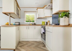 A compact kitchen with white cabinets, a herringbone floor, a window with blinds, and a small plant on the counter.