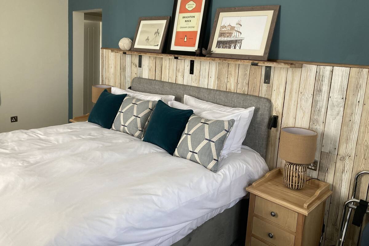 A neatly made bed with white linens and patterned pillows, a wooden nightstand with a lamp, and a wall-mounted shelf with framed pictures.