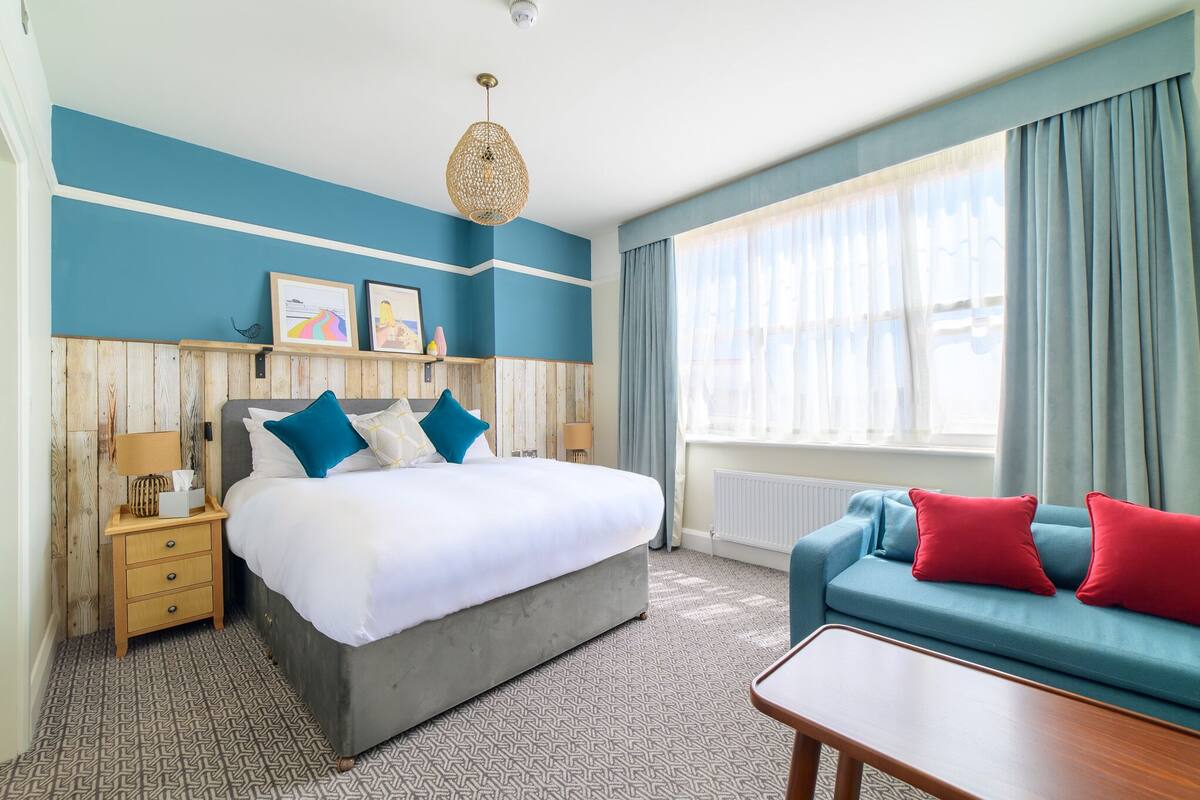 A hotel room with a large bed, a sofa, a wooden nightstand, and a hanging light fixture.