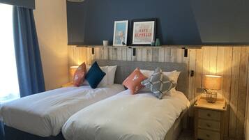 Una cama doble con ropa de cama blanca, dos mesitas de noche con lámparas, un cabecero de madera y una estantería en la pared con objetos decorativos.