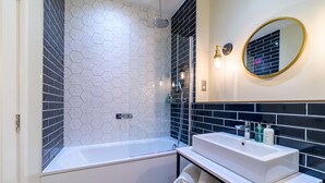 A modern bathroom with a hexagonal tile wall, a white bathtub, a white sink with a mirror, and a towel rack. - Downs Hotel (Brighton)