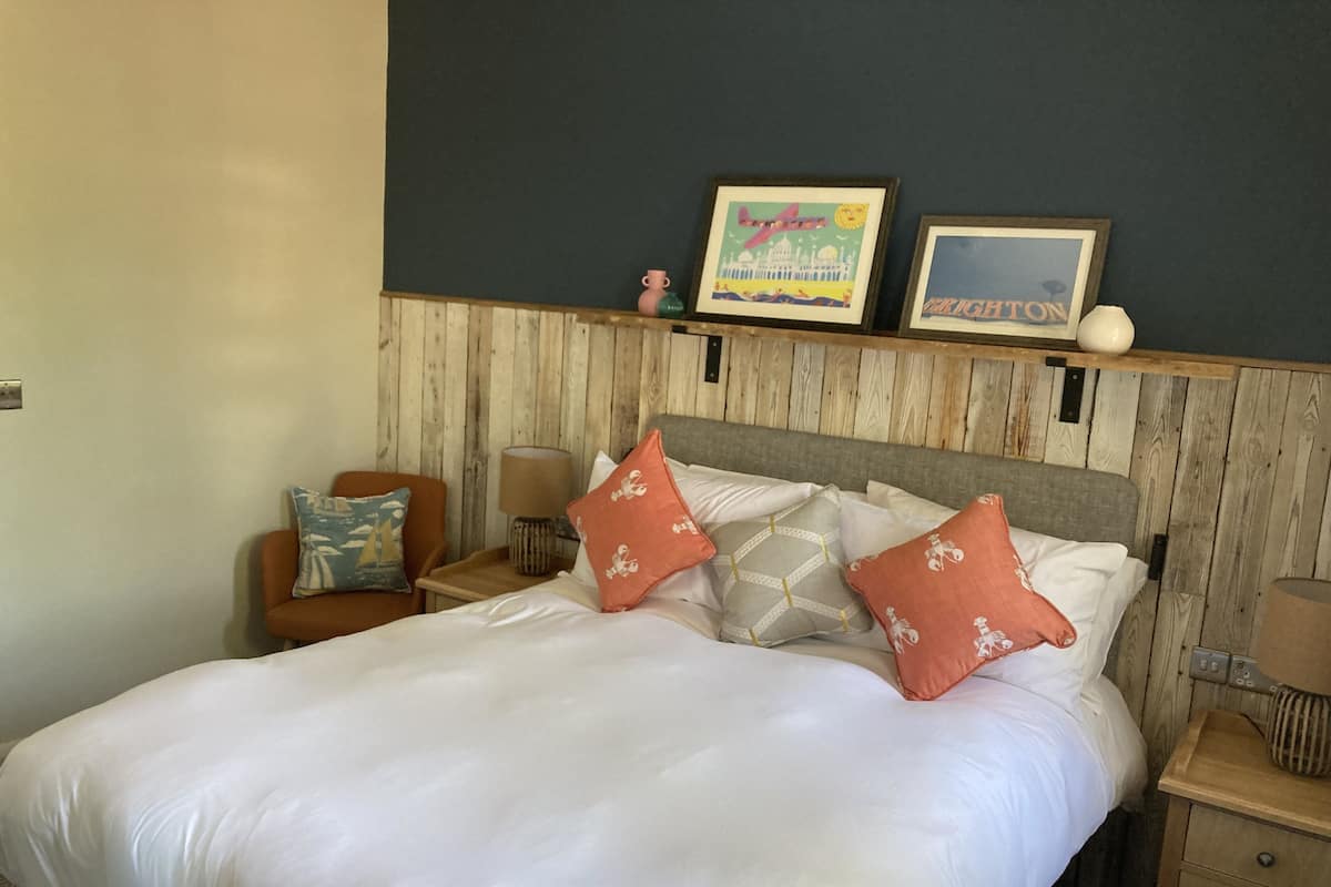 A neatly made bed with white linens and multiple decorative pillows, a wooden headboard, a small wooden nightstand with a lamp, and two framed pictures on the wall above the bed.