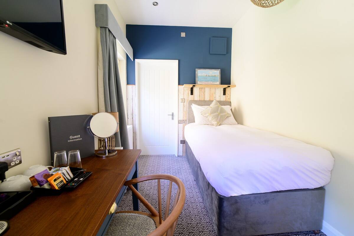A hotel room with a bed, a desk with a computer and a telephone, a mirror, and a door leading to the bathroom.