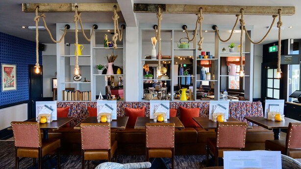 A restaurant with wooden tables and chairs, hanging rope lights, and shelves with various items. - Downs Hotel (Brighton)