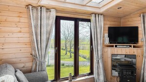 L’intérieur d’un chalet accueillant, avec un canapé, une cheminée, un téléviseur et une grande fenêtre agrémentée de rideaux.