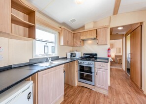 A compact kitchen with wooden cabinets, a built-in oven, a microwave, and a sink. - 12ft-wide caravan with one double and one twin. (Bognor Regis)