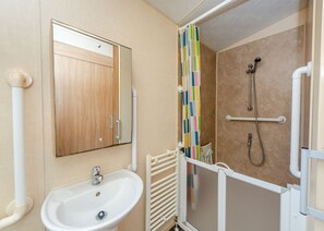 Ein Badezimmer mit Holztür, Waschbecken in Weiß, einer Dusche mit Haltegriff und einem gefliesten Duschbereich.