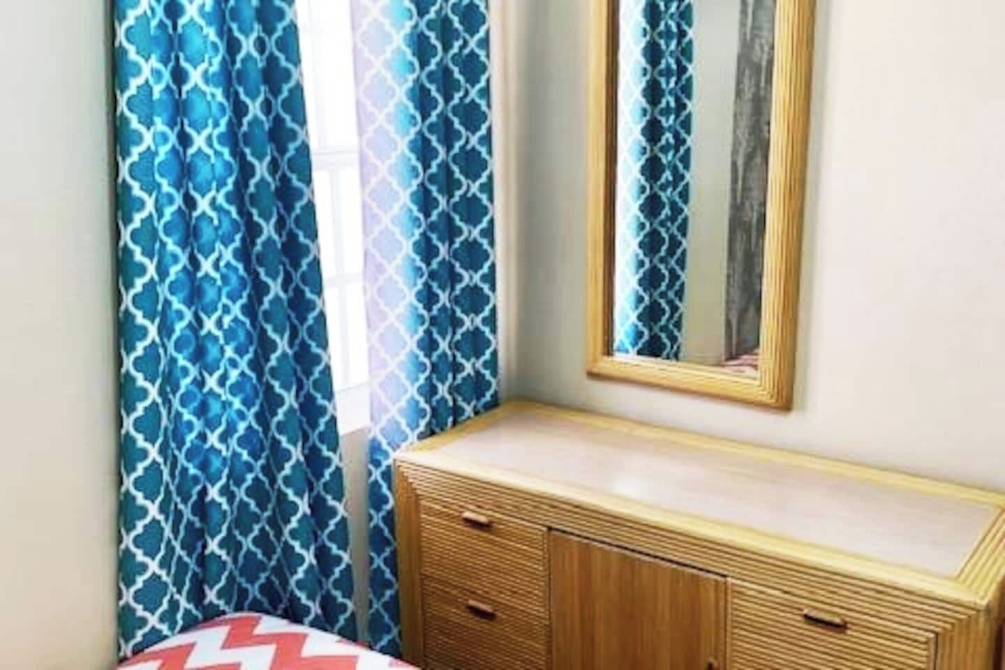 Ein Zimmer mit einem Bett, einem Nachttisch, einem Spiegel und einem Fenster mit blauen Vorhängen mit Muster.