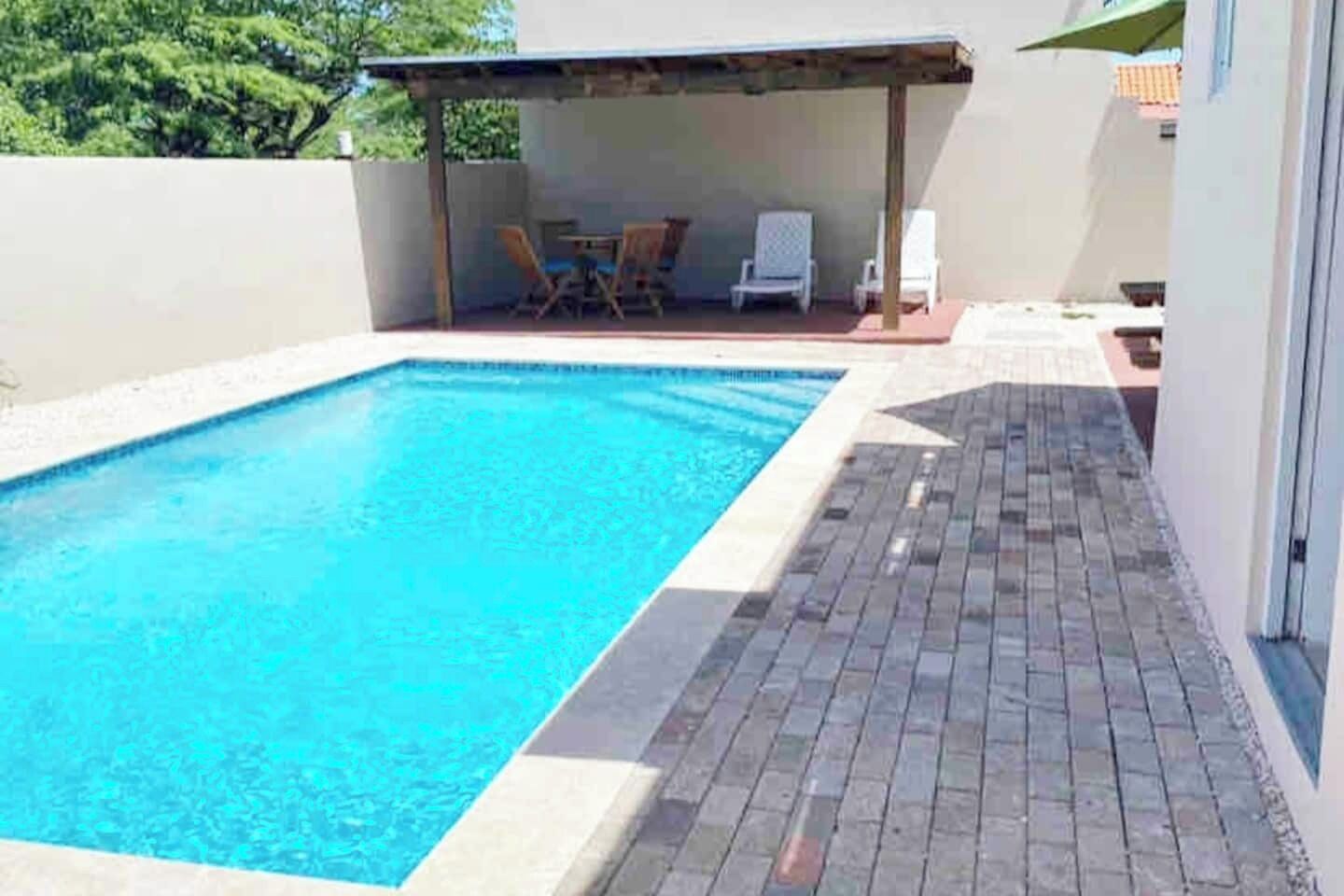 Ein rechteckiger Pool mit gefliestem Deckbereich, einem überdachten Sitzbereich mit Stühlen und Blick auf Bäume im Hintergrund.