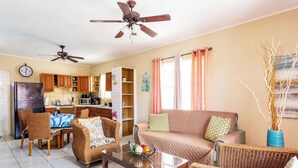Ein Wohnzimmer mit Korbmöbeln, einem Glastisch, einer Couch und einem Essbereich mit Tisch und Stühlen. Es gibt Deckenventilatoren, eine Uhr und ein Fenster mit Vorhängen.