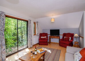 Un salon avec un téléviseur, deux fauteuils rouges, une table basse en bois avec des verres à vin et des livres, et une grande fenêtre avec des rideaux.