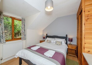 Une chambre avec un plafond en pente, un lit avec une literie blanche, deux lampes de chevet, une fenêtre avec des rideaux et un radiateur.