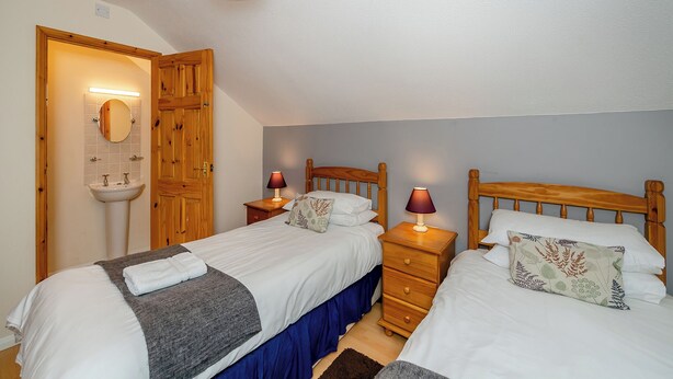 Een slaapkamer met twee bedden, een houten garderobe, een nachtkastje met een lamp en een badkamer met een wastafel en spiegel.