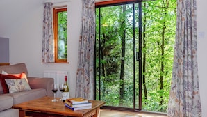 Un soggiorno con divano, tavolino con libri e bicchieri da vino, e una porta scorrevole in vetro che dà su una zona verde esterna.