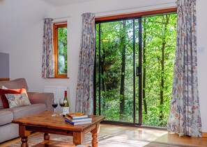 A living room with a sofa, a coffee table with books and wine glasses, and a sliding glass door leading to a green outdoor area.